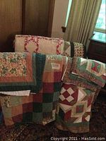 Multiple folded queen-sized quilts with various patchwork and floral patterns draped over furniture.