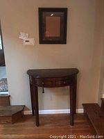 Photo showing the full wooden demilune table against a beige wall, hardwood floor, flanked by stairs.