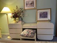 Cream-colored laminate dresser vanity with six drawers, rounded edges, and wooden trim handles, paired with a matching bench and floral cushion, shown in a bedroom setting with artwork and lamp.