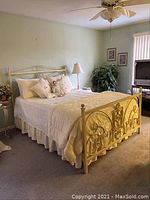 View of queen size bed dressed with cream bedding and decorative pillows and doll on the bed, showing metal headboard.