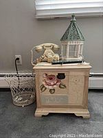 Photo showing storage cube with floral front panel, vintage phone, decorative bird cage, alarm clock and wire basket set against a wall with baseboard heater.