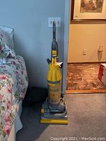 Full view of the upright Dyson vacuum cleaner with yellow cyclone chamber and gray base next to a bed on carpeted floor.