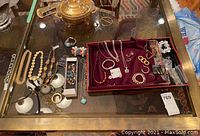 Jewelry collection displayed on glass table including gold-toned chain necklaces and bracelets, assortment of costume jewelry pieces in red velvet tray.