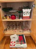 View of wooden cabinet shelf with Spode Christmas ceramic platter, glassware with Christmas tree designs, assorted Christmas ornaments, decorative green serving dish, and fabric napkins.