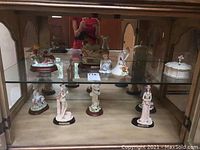 Two shelves of Lenox figurines and a white trinket box with gold accents on glass shelves inside a display cabinet