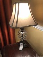 View of one lamp illuminated showing fabric shade with dark trim and faceted glass twist base on a wooden table.