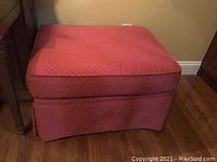 Full view of the red cloth upholstered ottoman with diamond pattern fabric and skirted bottom.