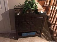 Front view of brown patterned cabinet with closed doors, bottom open shelf with blue box, and various items on top including boxes and plant.