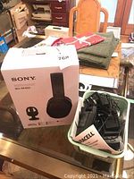 Sony WH-RF400 wireless headphones original box on a table with some folded cloth and a basket of chargers and adapters next to it.