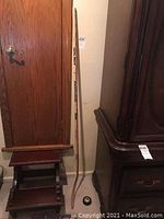 Long view of wooden hockey stick leaning against wall next to small wooden step stool and dark wooden cabinet, hockey puck on floor.