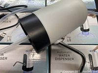 Close-up of one automatic water dispenser showing white body with black base and attached clear plastic tubing and USB power cord resting on boxes.