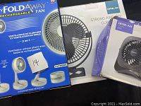 Three boxed foldaway and clip-on fans in original packaging. Visible brand names and model information on boxes.