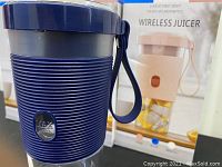 Front view of one dark blue wireless juicer with ribbed body and strap handle with product box in background