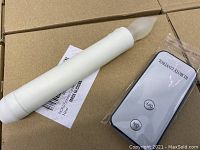 White flameless LED candle with frosted flame tip placed on cardboard surface beside a sealed remote control.