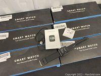 Set of six black smart watches with black silicone wristbands, smart watch boxes, charging cables, and instruction manuals on display.