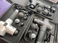 Photo showing all three massage guns in their respective cases along with multiple massage head attachments and charging cables.