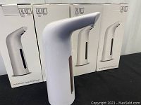 Three automatic soap dispensers displayed with their original packaging boxes in the background. The dispenser in front is white with a window showing soap level.