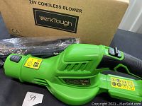 Green WerkTough 20V cordless blower, side view with black grip and power switch, yellow warning labels, and nozzle attachments wrapped in plastic, placed beside the box.