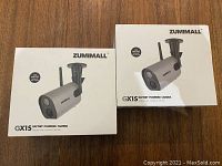 Two Zumimall GX1S battery powered camera boxes on a wood surface, showing product packaging and brand