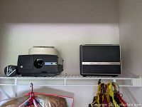 Photo showing the Bell & Howell slide projector with carousel and a camera lens on the left side and the Bell & Howell Puriosound sound system unit on the right side of a wire shelf.