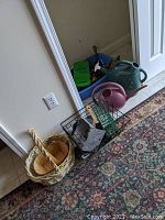 Photo showing woven baskets, metal baskets, plastic watering cans, and assorted gardening tools placed on the floor near a door.