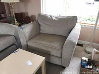 Oversized chair with textured light beige fabric upholstery showing armrest and back cushion with some wear, positioned next to a beige table and adjacent sofa.