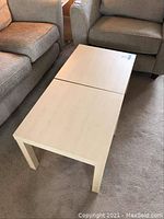 Photo showing two light wood finish square IKEA tables set next to each other between armchairs on a carpeted floor.