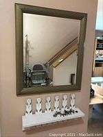 Full view of the wall mirror, white floral shelf below with small items on top; part of the metal coatrack visible with umbrellas hanging.