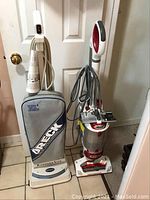 Oreck and Shark upright vacuum cleaners side by side in front of a door.