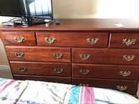 Full front view of the dark wood dresser showing all nine drawers and brass handles, with TV and remote on top.
