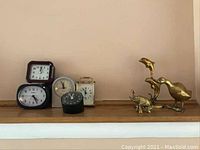 Photo shows five different alarm clocks arranged on a wooden shelf alongside three brass animal figurines including a dolphin, an elephant, and a bird. Various styles and sizes shown.