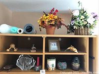 View of faux floral arrangements, figurines, and candles displayed on shelves. Shows autumn sunflower arrangement and smaller figurines below.