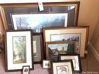 Multiple framed artwork in various sizes, leaning against a furniture piece in an indoor setting. Scenes include nature landscapes, hunting, and wildlife.
