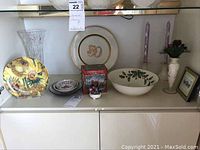 Objects arranged on white cabinet shelf: crystal vase, sunflower plate, Lenox Christmas bowl, porcelain plates, candle holders, small framed artwork, flower candle holder.