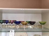 Full shelf of 12 colorful cut crystal glasses showing variety in colors and shapes, some glasses chipped.