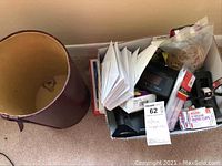 Wastepaper basket next to box filled with various office supplies including paper clips, mailing labels, adhesives, rubber bands, magnifying glass, and paper stacks.