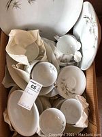 Box containing various cups, saucers, and plates wrapped in paper, showing some pieces with chips and the bamboo garden pattern.