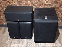 Subwoofer at right with three fabric-grille speakers stacked at left against wood panel wall.