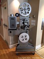 Full view of the vintage floor mounted 35mm film projector, showing metal body, large film reels and base with wheels.