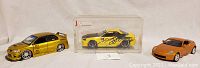 Three diecast model cars shown together: gold Nissan Skyline GTR V-SPEC II on left, yellow Mitsubishi Lancer Evolution in center display case, orange Nissan 350Z on right.