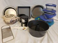 Photo showing assorted kitchen items including metal baking pans, a ceramic teapot, a handheld metal grater, a ceramic Disney spoon rest, several covered containers stacked, and a round tube baking pan.
