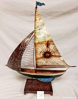 Model decorative sailboat with artistic metal sails, wooden hull painted white and brown, mounted on a rectangular base with script pattern. Shows a blue flag on the mast.