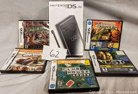 Photo showing Nintendo DS Lite original box, five Nintendo DS game cases arranged behind the box with visible cover art, and seller's lot number card.