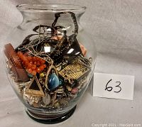 Full clear glass vase filled with various pieces of costume jewelry in different colors and materials, showing multiple bracelets, necklaces, and earrings.
