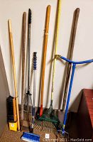 Full view of gardening tools propped against wall showing shovel, two leaf rakes, claw, broom, and brush.