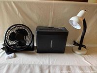 Photo shows black 8-inch Home Design desktop fan, black Embassy paper shredder, and white gooseneck desk lamp on a beige cloth background.