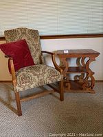 Full view of the tapestry upholstered Fairfield armchair with red bead fringe accent pillow and knotty pine end table beside it