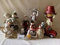 Multiple whimsical snowmen figurines of various sizes grouped together, including one snow angel and a toy red car, arranged on a flat surface against a beige wall.