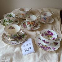 Full set of six tea cups and six matching saucers arranged on cream cloth, showing various floral patterns and gold trim.