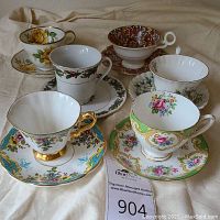 Front view of six bone china teacups and matching saucers displaying varied floral and gold trim designs on cream fabric background.